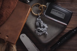 A collection of personal items arranged on a wooden surface, including a leather-bound notebook, a metal keychain with a dragon design, a car key for a Jaguar, a striped pocket knife, and a tactical pen.