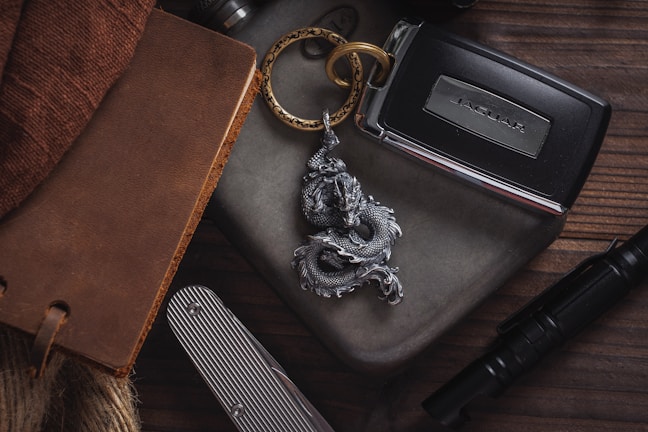 A collection of personal items arranged on a wooden surface, including a leather-bound notebook, a metal keychain with a dragon design, a car key for a Jaguar, a striped pocket knife, and a tactical pen.