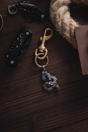 A wooden surface featuring a brass hook with intricate dragon detailing, two black folding knives with visible screws and openings, a thick coiled rope, and the corner of a brown leather-bound book.