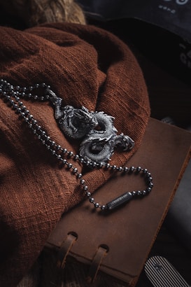 A textured brown fabric is draped over a leather-bound journal. A black beaded necklace featuring an intricately detailed dragon pendant is artfully arranged on top. The setting suggests a rustic or vintage theme.