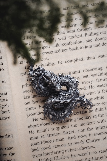A detailed metallic dragon sculpture rests on the pages of an open book with visible text. The sculpture has intricate scales and a classic dragon form with curves and fierce details. Shadows of leaves or foliage fall across the page, adding depth and texture to the scene.