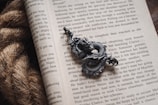 A detailed close-up of an open book with text visible on its pages, accompanied by a dark metallic dragon ornament placed on top of the pages. The texture of the dragon is intricate, with scales and curves giving it a lifelike appearance. Beside the book, a coil of rough, natural-colored rope is partially visible.