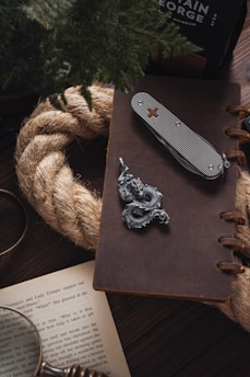 A rustic setting featuring a leather-bound notebook placed on a wooden surface, accompanied by a silver dragon pendant and a multi-tool with a cross emblem. A thick rope is coiled next to an open book with visible text, and partially obscured greenery adds a natural element.