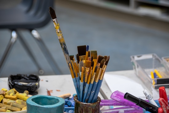 Various paintbrushes of different sizes and shapes are arranged in a holder on a cluttered art desk. Other art supplies, including chalk pastels, markers, and containers, are scattered around, with a classroom chair and shelving visible in the background.