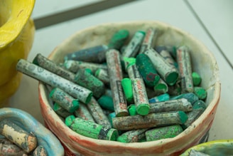 A collection of colorful oil pastels are displayed in a ceramic bowl. The pastels are mostly shades of green and gray, with visible markings and labels on some pieces. Adjacent to the bowl is a bright yellow ceramic vase, adding a pop of color to the scene.