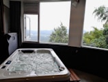 A luxurious hot tub sits indoors near a large window, offering a view of a scenic landscape with lush green trees and distant hills under a cloudy sky.