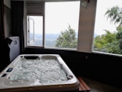 A luxurious hot tub sits indoors near a large window, offering a view of a scenic landscape with lush green trees and distant hills under a cloudy sky.