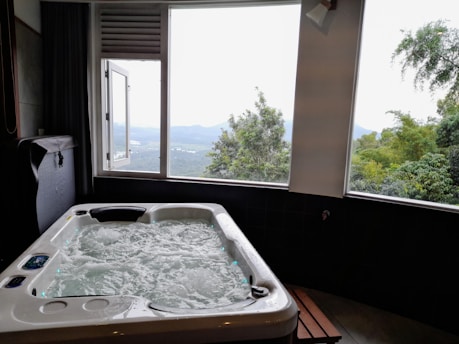 A luxurious hot tub sits indoors near a large window, offering a view of a scenic landscape with lush green trees and distant hills under a cloudy sky.