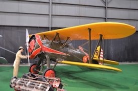 A vintage biplane with bright yellow wings and red accents is displayed indoors. A mannequin dressed in period clothing stands near the propeller, suggesting a historical aviation scene. The aircraft is set against a dark backdrop, and an engine rests on the floor in the foreground.