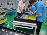 Three people are assembling electronics in a manufacturing facility. They are working with a rectangular electronic component on a white table. The background includes blue pallets and industrial shelving. The floor is green with yellow and black hazard markings.