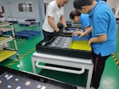 Three people are assembling electronics in a manufacturing facility. They are working with a rectangular electronic component on a white table. The background includes blue pallets and industrial shelving. The floor is green with yellow and black hazard markings.