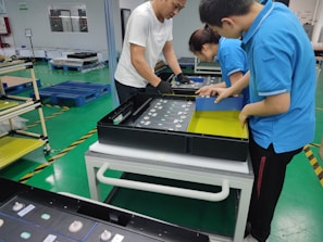 Three people are assembling electronics in a manufacturing facility. They are working with a rectangular electronic component on a white table. The background includes blue pallets and industrial shelving. The floor is green with yellow and black hazard markings.