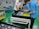 Three people are assembling electronics in a manufacturing facility. They are working with a rectangular electronic component on a white table. The background includes blue pallets and industrial shelving. The floor is green with yellow and black hazard markings.