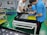 Three people are assembling electronics in a manufacturing facility. They are working with a rectangular electronic component on a white table. The background includes blue pallets and industrial shelving. The floor is green with yellow and black hazard markings.