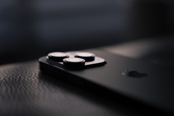 A close-up view of a sleek, modern smartphone lying on a textured surface. The focus is on the camera lenses, with the rest of the phone fading into the background, emphasizing its minimalist design.