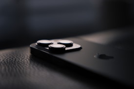 A close-up view of a sleek, modern smartphone lying on a textured surface. The focus is on the camera lenses, with the rest of the phone fading into the background, emphasizing its minimalist design.