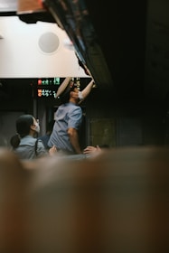 A person wearing a uniform and cap is reaching upwards inside a vehicle or a train, with their arm extended towards some equipment or luggage compartment. Another individual, also in uniform and wearing a face mask, is assisting or observing. The setting is dimly lit, and there are electronic signs displaying alphanumeric characters in the background.