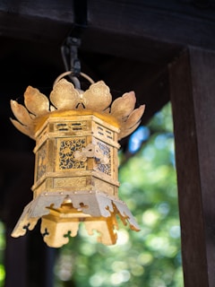 A intricately designed, golden hanging lantern with floral and geometric patterns. The lantern is ornate, featuring decorative metalwork with a slightly aged patina. It is suspended under a wooden structure with soft, natural light creating a blurred green and blue bokeh in the background.