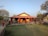 A small bungalow with a red-tiled roof and yellow walls is surrounded by a garden area. The house features a porch with decorative red pillars and outdoor seating. The landscape includes trees and patches of grass, situated under a clear blue sky.