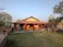 A small bungalow with a red-tiled roof and yellow walls is surrounded by a garden area. The house features a porch with decorative red pillars and outdoor seating. The landscape includes trees and patches of grass, situated under a clear blue sky.