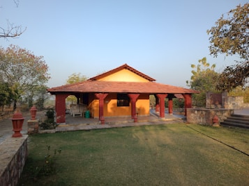 A small bungalow with a red-tiled roof and yellow walls is surrounded by a garden area. The house features a porch with decorative red pillars and outdoor seating. The landscape includes trees and patches of grass, situated under a clear blue sky.