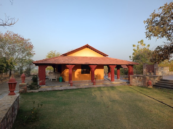 A small bungalow with a red-tiled roof and yellow walls is surrounded by a garden area. The house features a porch with decorative red pillars and outdoor seating. The landscape includes trees and patches of grass, situated under a clear blue sky.
