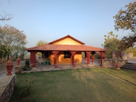 A small bungalow with a red-tiled roof and yellow walls is surrounded by a garden area. The house features a porch with decorative red pillars and outdoor seating. The landscape includes trees and patches of grass, situated under a clear blue sky.
