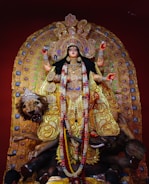 A richly adorned statue of a goddess with multiple arms, wearing a lavish gold and jewel-studded dress. She is surrounded by intricate decorations and stands atop a lion. The statue is detailed with flowers and vibrant patterns, displaying a sense of grandeur and divinity.