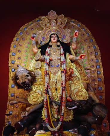 A richly adorned statue of a goddess with multiple arms, wearing a lavish gold and jewel-studded dress. She is surrounded by intricate decorations and stands atop a lion. The statue is detailed with flowers and vibrant patterns, displaying a sense of grandeur and divinity.