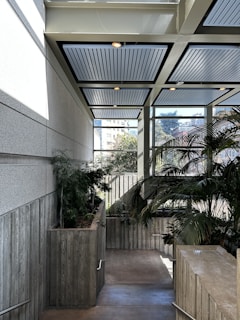 An interior space featuring a staircase with wooden planter boxes lining one side, filled with lush green plants. Large windows provide a view of an urban landscape with buildings and trees, allowing natural light to flood the area. The ceiling is modern with recessed lighting and paneling.