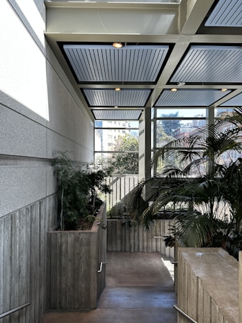 An interior space featuring a staircase with wooden planter boxes lining one side, filled with lush green plants. Large windows provide a view of an urban landscape with buildings and trees, allowing natural light to flood the area. The ceiling is modern with recessed lighting and paneling.