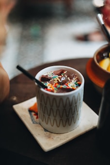 A ceramic mug decorated with wavy patterns is filled with a rich chocolate drink topped with colorful sprinkles. The mug is placed on a square tile coaster and has a black straw inside.