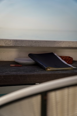 A slightly blurred image featuring a wooden surface with an assortment of objects. A closed black menu or book rests atop white plates, accompanied by a small rectangular object, possibly a card. Off to the side, part of a woven basket is partially visible.