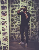 A person stands in a small room with walls completely covered in a collage of black-and-white photographs. The person is holding a camera up to their face, partially obscuring their features, and is dressed casually in a dark top and jeans. They are wearing stylish sneakers with colorful accents and a watch. The overall atmosphere feels artistic and modern.