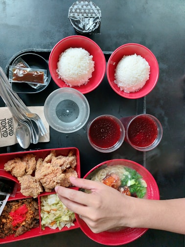 A variety of Asian dishes are arranged on a black table surface. There are two bowls of white rice in red bowls, a ramen dish with broth, noodles, and vegetables, and a bento box containing fried chicken, cabbage salad, minced meat with chili, and red pickled ginger. Two cups of red gelatin dessert and a plastic cup of water are alongside the food. Utensils and a napkin are placed on the side, and a hand is seen reaching towards the food.