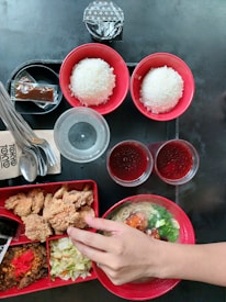 A variety of Asian dishes are arranged on a black table surface. There are two bowls of white rice in red bowls, a ramen dish with broth, noodles, and vegetables, and a bento box containing fried chicken, cabbage salad, minced meat with chili, and red pickled ginger. Two cups of red gelatin dessert and a plastic cup of water are alongside the food. Utensils and a napkin are placed on the side, and a hand is seen reaching towards the food.