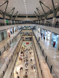 A spacious indoor shopping mall with multiple levels, featuring numerous shops lining the sides. There are people walking on each level, with some stopping to look into store windows. The architecture includes modern railings and a high ceiling with distinctive, geometric support beams. The floor is made of tiles in a neutral tone, and bright lighting illuminates the mall.
