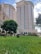 Two tall, modern residential buildings are surrounded by lush green grass and trees. The facade of the towers features a blend of light peach and cream colors, with numerous balconies. In the foreground, a well-maintained grassy area with scattered trees and flowering bushes adds to the urban landscape.