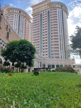 Two tall, modern residential buildings are surrounded by lush green grass and trees. The facade of the towers features a blend of light peach and cream colors, with numerous balconies. In the foreground, a well-maintained grassy area with scattered trees and flowering bushes adds to the urban landscape.