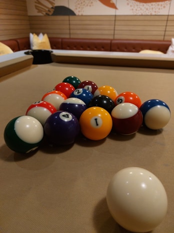 A set of billiard balls arranged on a pool table. The balls are colorful, featuring numbers, and the surrounding environment includes wooden panel walls and cushioned seating.