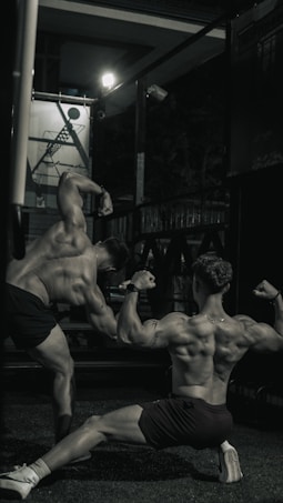 Two muscular individuals are flexing their back muscles in a dimly lit gym. They are positioned in a way to showcase their physique, with one kneeling on the ground and the other standing to the side. The environment appears industrial with gym equipment and a poster in the background.