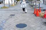 A person with short hair is walking away on a paved sidewalk, pulling a white suitcase and carrying a bag. They are dressed in casual clothing, including a gray hoodie and black pants. Several orange traffic cones line the path on the right, with some patches of greenery and trees visible around the pavement. In the background, a few other individuals are visible, walking in the distance near some buildings.