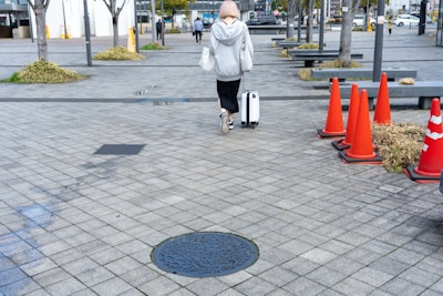 A person with short hair is walking away on a paved sidewalk, pulling a white suitcase and carrying a bag. They are dressed in casual clothing, including a gray hoodie and black pants. Several orange traffic cones line the path on the right, with some patches of greenery and trees visible around the pavement. In the background, a few other individuals are visible, walking in the distance near some buildings.