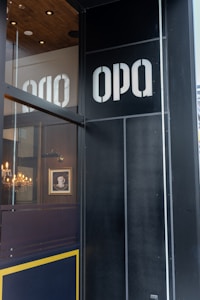 A storefront with large letters spelling 'OPA' on a dark, glass-panelled corner. Inside, a cozy interior features a chandelier and a framed picture of a steaming cup. The color scheme includes dark wood and gold accents, creating a warm atmosphere.