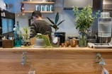 A cozy kitchen space features a wooden counter adorned with various potted plants, bottles, and decorative items. A man with short hair stands in the background, possibly preparing food. The ambiance is enhanced by the presence of natural elements and a warm, inviting atmosphere.