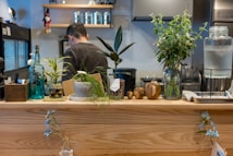 A cozy kitchen space features a wooden counter adorned with various potted plants, bottles, and decorative items. A man with short hair stands in the background, possibly preparing food. The ambiance is enhanced by the presence of natural elements and a warm, inviting atmosphere.