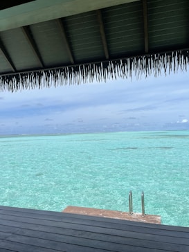 A serene view of a turquoise ocean stretches out to the horizon under a clear blue sky. A thatched roof partially covers the scene from above, casting a shadow over a wooden deck area. A small set of metal steps leads down to the inviting water.