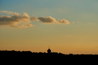 A silhouette of a domed structure with a cross at the top appears against the backdrop of an orange and blue sky. Fluffy clouds float above the horizon, creating a serene atmosphere in the twilight.