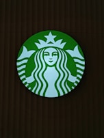 A round green logo with a white silhouette of a twin-tailed siren in the center. The siren has long wavy hair, a serene facial expression, and is framed by a circular band with a star above her head.