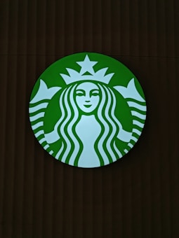 A round green logo with a white silhouette of a twin-tailed siren in the center. The siren has long wavy hair, a serene facial expression, and is framed by a circular band with a star above her head.